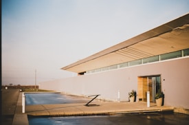 A modern, minimalist building with clean lines and a flat roof is presented under a clear sky. The structure has large glass windows and light-colored walls. There is a concrete patio with a reflective water feature in front, along with several potted plants. The setting appears tranquil and spacious, hinting at a suburban or rural location.