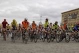 A large group of cyclists, wearing helmets and colorful clothing, is riding together on a cobblestone street. Many cyclists are wearing bright orange safety vests. In the background, there is a large building with a sign that reads 'DAO Investment Group' and a cloudy sky.