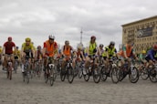 A large group of cyclists, wearing helmets and colorful clothing, is riding together on a cobblestone street. Many cyclists are wearing bright orange safety vests. In the background, there is a large building with a sign that reads 'DAO Investment Group' and a cloudy sky.