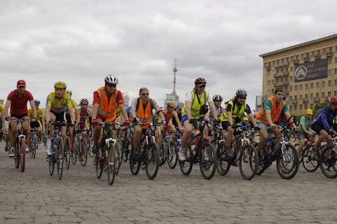 A large group of cyclists, wearing helmets and colorful clothing, is riding together on a cobblestone street. Many cyclists are wearing bright orange safety vests. In the background, there is a large building with a sign that reads 'DAO Investment Group' and a cloudy sky.