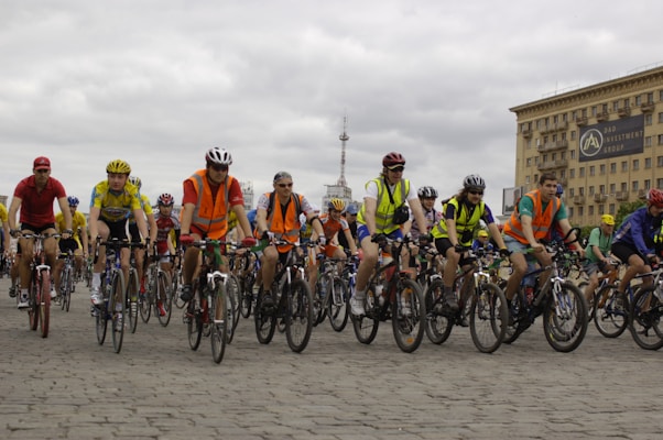 A large group of cyclists, wearing helmets and colorful clothing, is riding together on a cobblestone street. Many cyclists are wearing bright orange safety vests. In the background, there is a large building with a sign that reads 'DAO Investment Group' and a cloudy sky.