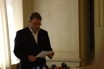 A man wearing a formal suit is reading a piece of paper while standing in a room with soft lighting. The background features a wall with ornate details and a partially open curtain letting in light. There are microphones and bottles in front of him, suggesting a possible press conference or formal event.