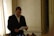 A man wearing a formal suit is reading a piece of paper while standing in a room with soft lighting. The background features a wall with ornate details and a partially open curtain letting in light. There are microphones and bottles in front of him, suggesting a possible press conference or formal event.