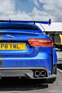 A vibrant blue sports car is parked, showcasing its rear with a large spoiler and dual exhaust pipes. The car has a prominent Jaguar emblem and a yellow license plate. The sky is partially cloudy, and there are tents in the background, suggesting an outdoor setting, possibly a car event. A person wearing a yellow outfit is visible in the background.