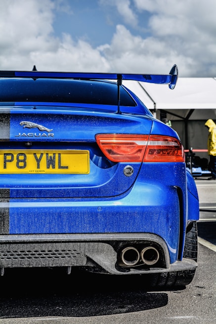 A vibrant blue sports car is parked, showcasing its rear with a large spoiler and dual exhaust pipes. The car has a prominent Jaguar emblem and a yellow license plate. The sky is partially cloudy, and there are tents in the background, suggesting an outdoor setting, possibly a car event. A person wearing a yellow outfit is visible in the background.