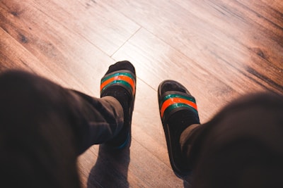 A pair of feet wearing black socks and striped slip-on sandals with green and orange bands, positioned on a wooden floor.