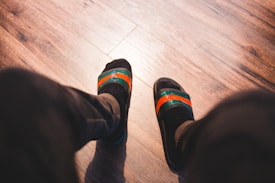 A pair of feet wearing black socks and striped slip-on sandals with green and orange bands, positioned on a wooden floor.