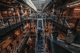A multi-level shopping mall interior with people riding escalators and walking around. The image includes a variety of shops on both levels, with escalators in the center. The floors have a geometric checkered pattern, and there is ambient lighting from the ceiling. Shoppers, some carrying shopping bags, create a busy and lively atmosphere.