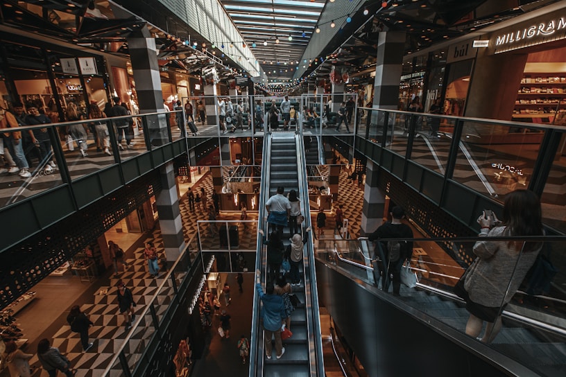 A multi-level shopping mall interior with people riding escalators and walking around. The image includes a variety of shops on both levels, with escalators in the center. The floors have a geometric checkered pattern, and there is ambient lighting from the ceiling. Shoppers, some carrying shopping bags, create a busy and lively atmosphere.
