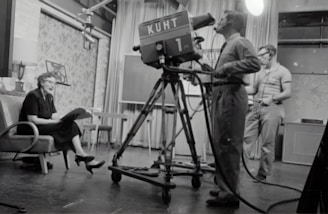 grayscale photography of man standing near studio camera and woman sitting while holding book
