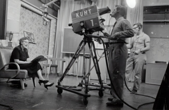 grayscale photography of man standing near studio camera and woman sitting while holding book