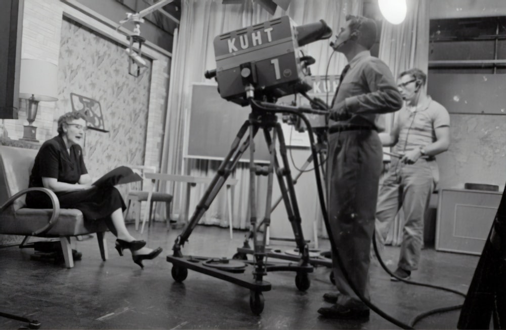 스튜디오 카메라 근처에 서 있는 남자와 책을 들고 앉아 있는 여자의 회색조 사진
