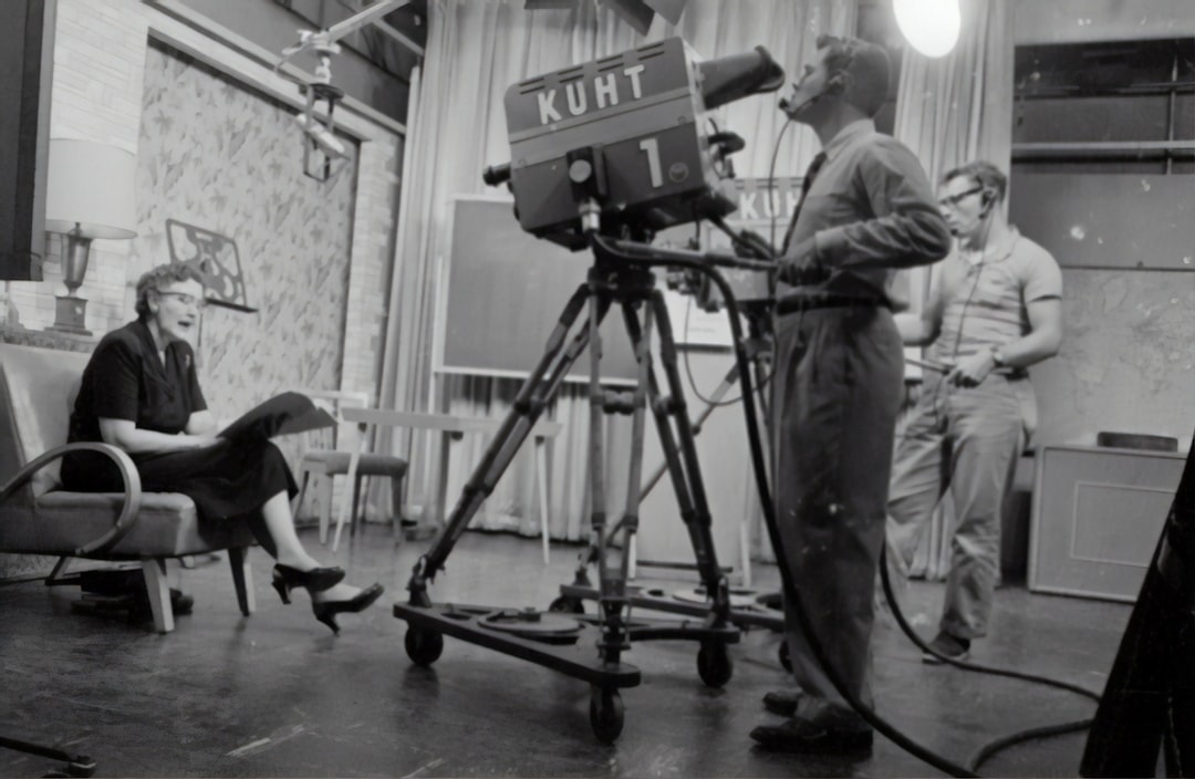 fotografía en escala de grises de un hombre de pie cerca de una cámara de estudio y una mujer sentada sosteniendo un libro