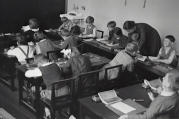 A group of young children sits at wooden desks in a classroom setting, with a teacher assisting them. The children are engaged in writing or drawing activities, wearing clothing typical of an earlier era. The teacher leans over to help some of the children, indicating an interactive and attentive learning environment. The room has a minimalist design with a blackboard and simple furniture.