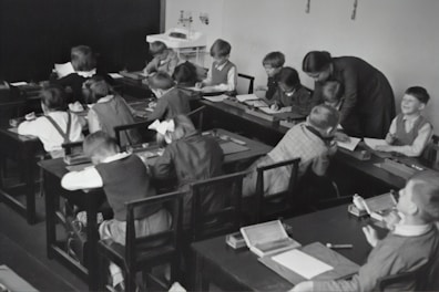 A group of young children sits at wooden desks in a classroom setting, with a teacher assisting them. The children are engaged in writing or drawing activities, wearing clothing typical of an earlier era. The teacher leans over to help some of the children, indicating an interactive and attentive learning environment. The room has a minimalist design with a blackboard and simple furniture.
