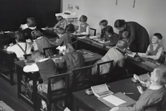 A group of young children sits at wooden desks in a classroom setting, with a teacher assisting them. The children are engaged in writing or drawing activities, wearing clothing typical of an earlier era. The teacher leans over to help some of the children, indicating an interactive and attentive learning environment. The room has a minimalist design with a blackboard and simple furniture.