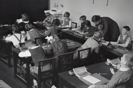A group of young children sits at wooden desks in a classroom setting, with a teacher assisting them. The children are engaged in writing or drawing activities, wearing clothing typical of an earlier era. The teacher leans over to help some of the children, indicating an interactive and attentive learning environment. The room has a minimalist design with a blackboard and simple furniture.