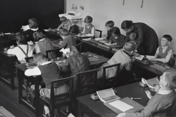 A group of young children sits at wooden desks in a classroom setting, with a teacher assisting them. The children are engaged in writing or drawing activities, wearing clothing typical of an earlier era. The teacher leans over to help some of the children, indicating an interactive and attentive learning environment. The room has a minimalist design with a blackboard and simple furniture.