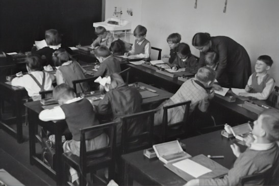 A group of young children sits at wooden desks in a classroom setting, with a teacher assisting them. The children are engaged in writing or drawing activities, wearing clothing typical of an earlier era. The teacher leans over to help some of the children, indicating an interactive and attentive learning environment. The room has a minimalist design with a blackboard and simple furniture.
