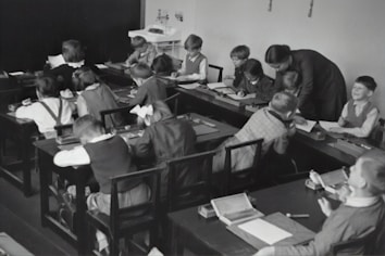 A group of young children sits at wooden desks in a classroom setting, with a teacher assisting them. The children are engaged in writing or drawing activities, wearing clothing typical of an earlier era. The teacher leans over to help some of the children, indicating an interactive and attentive learning environment. The room has a minimalist design with a blackboard and simple furniture.
