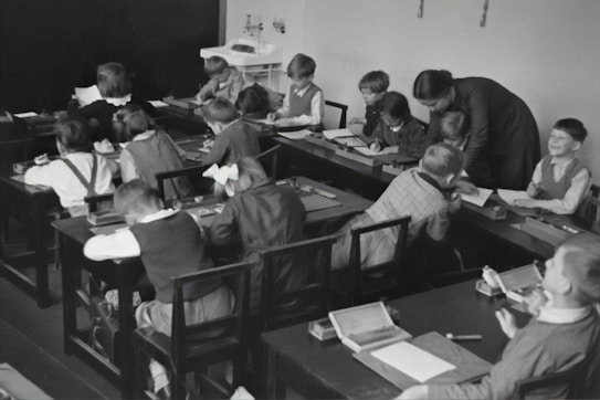 A group of young children sits at wooden desks in a classroom setting, with a teacher assisting them. The children are engaged in writing or drawing activities, wearing clothing typical of an earlier era. The teacher leans over to help some of the children, indicating an interactive and attentive learning environment. The room has a minimalist design with a blackboard and simple furniture.
