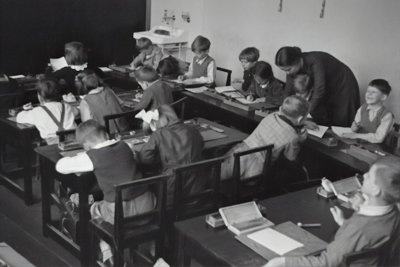 A group of young children sits at wooden desks in a classroom setting, with a teacher assisting them. The children are engaged in writing or drawing activities, wearing clothing typical of an earlier era. The teacher leans over to help some of the children, indicating an interactive and attentive learning environment. The room has a minimalist design with a blackboard and simple furniture.