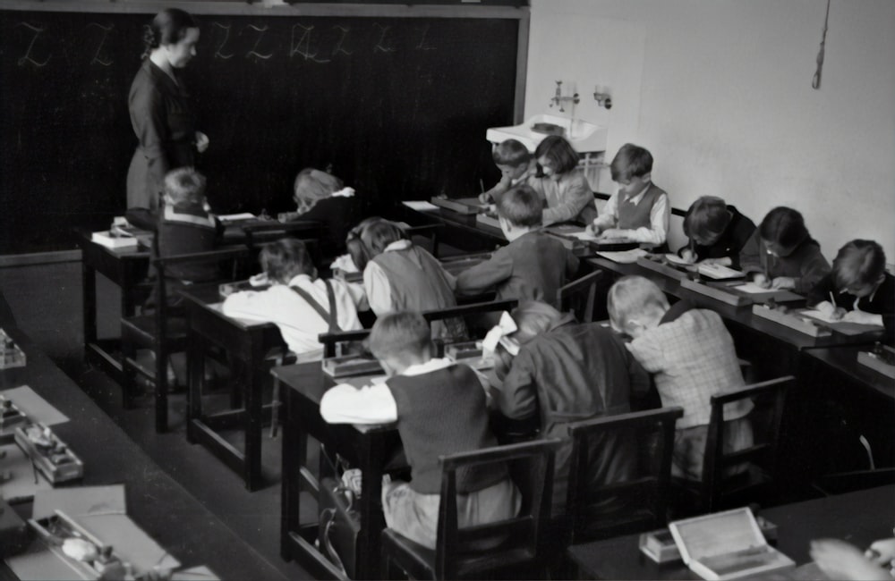 黒板のそばに立つ教師と椅子に座る子供たちのグレースケール写真