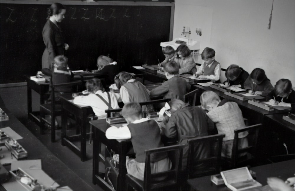 grayscale photography of teacher standing near chalkboard and children sitting on chairs