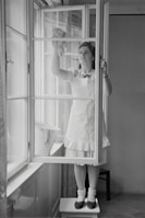 A woman in a dress and apron stands on a small table, cleaning a window with a cloth. The setting is indoors, with light streaming through large windows. A bucket is placed on the window sill nearby.