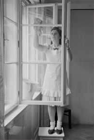 A woman in a dress and apron stands on a small table, cleaning a window with a cloth. The setting is indoors, with light streaming through large windows. A bucket is placed on the window sill nearby.