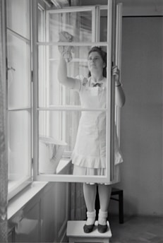 A woman in a dress and apron stands on a small table, cleaning a window with a cloth. The setting is indoors, with light streaming through large windows. A bucket is placed on the window sill nearby.