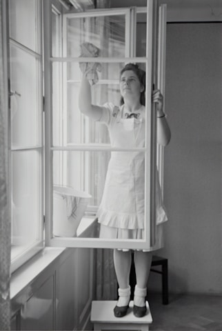 A woman in a dress and apron stands on a small table, cleaning a window with a cloth. The setting is indoors, with light streaming through large windows. A bucket is placed on the window sill nearby.