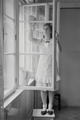 A woman in a dress and apron stands on a small table, cleaning a window with a cloth. The setting is indoors, with light streaming through large windows. A bucket is placed on the window sill nearby.