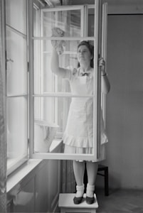 A woman in a dress and apron stands on a small table, cleaning a window with a cloth. The setting is indoors, with light streaming through large windows. A bucket is placed on the window sill nearby.