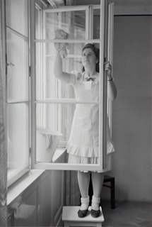 A woman in a dress and apron stands on a small table, cleaning a window with a cloth. The setting is indoors, with light streaming through large windows. A bucket is placed on the window sill nearby.