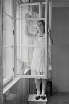 A woman in a dress and apron stands on a small table, cleaning a window with a cloth. The setting is indoors, with light streaming through large windows. A bucket is placed on the window sill nearby.