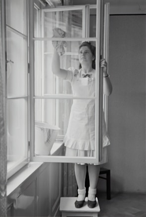 A woman in a dress and apron stands on a small table, cleaning a window with a cloth. The setting is indoors, with light streaming through large windows. A bucket is placed on the window sill nearby.