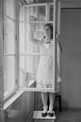 A woman in a dress and apron stands on a small table, cleaning a window with a cloth. The setting is indoors, with light streaming through large windows. A bucket is placed on the window sill nearby.