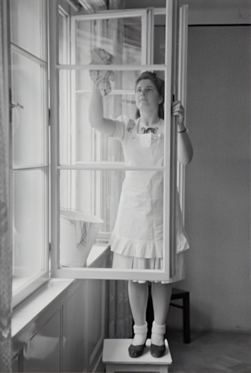 A woman in a dress and apron stands on a small table, cleaning a window with a cloth. The setting is indoors, with light streaming through large windows. A bucket is placed on the window sill nearby.