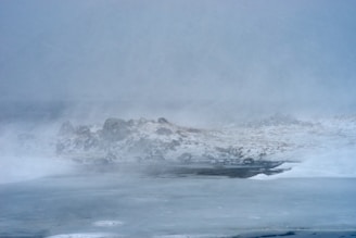 A desolate winter landscape under heavy fog, with snow-covered ground and rocky outcrops partially visible. The scene evokes a sense of cold and isolation.