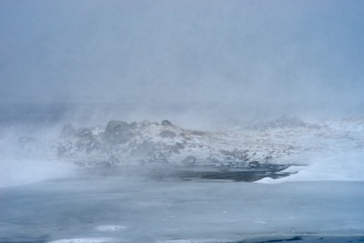 A desolate winter landscape under heavy fog, with snow-covered ground and rocky outcrops partially visible. The scene evokes a sense of cold and isolation.