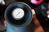 A close-up of a black kettle with a thermometer on top displaying temperature in degrees Celsius and Fahrenheit. The kettle handle is visible, and the focus is on the clearly marked thermometer dial.