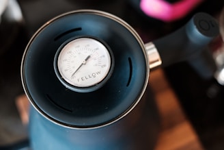 A close-up of a black kettle with a thermometer on top displaying temperature in degrees Celsius and Fahrenheit. The kettle handle is visible, and the focus is on the clearly marked thermometer dial.