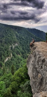 A person sits on the edge of a high cliff overlooking a vast, dense forest under a cloudy sky. The scene conveys a sense of adventure and solitude.