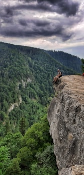 A person sits on the edge of a high cliff overlooking a vast, dense forest under a cloudy sky. The scene conveys a sense of adventure and solitude.