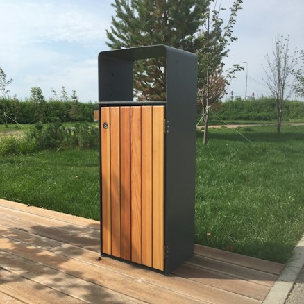 A modern outdoor trash can with a wooden front panel and a sleek black metal frame is situated on a wooden pathway. It is surrounded by green grass and nearby trees, under a partly cloudy sky.