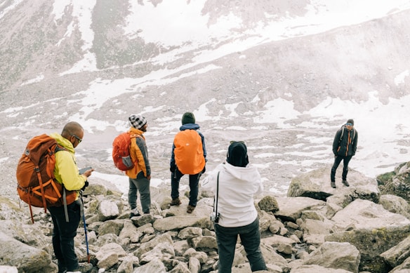 A group of people wearing outdoor gear stand on rocky terrain with snow scattered around. Some individuals have backpacks, and one person is using hiking poles. In the background, a large, snow-covered mountain slope is visible.