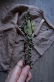 A hand holding a bundle of dried herbs wrapped together with twine. The bundle includes green leaves and small purple flowers. It rests against a wrinkled fabric background and a piece of wood is visible lying on the fabric.