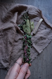 A hand holding a bundle of dried herbs wrapped together with twine. The bundle includes green leaves and small purple flowers. It rests against a wrinkled fabric background and a piece of wood is visible lying on the fabric.