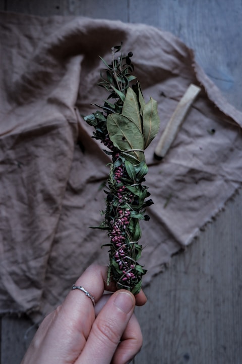 A hand holding a bundle of dried herbs wrapped together with twine. The bundle includes green leaves and small purple flowers. It rests against a wrinkled fabric background and a piece of wood is visible lying on the fabric.